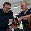 Wolfgang Lex (rechts) mit einem fahrerlosen Transportsystem von Fischertechnik. Dieses orientiert sich mit Ultraschallsensoren, Liniensensoren, Tastern und einem Fototransistor innerhalb der Agile Production Simulation.
