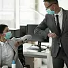 Eine Frau und ein Mann, die beide eine Maske tragen, klatschen sich in einem Büro mit dem Ellenbogen ab.