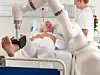 Eine Pflegerin spricht mit einem Patienten, der im Bett liegt. An seinem Fuß arbeitet ein Reha-Roboter.
