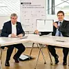 Holger Hanselka (links), Präsident KIT, und Heinz-Jürgen Prokop, CEO Trumpf Werkzeugmaschinen, sitzen an einem Tisch. Prokop hält den Kooperationsvertrag in die Höhe.