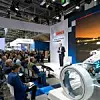 Ein Sprecher von Bosch präsentiert auf einer Messebühne vor einem großen Publikum. Auf der Bühne steht er neben einer Leinwand mit einer Projektion eines Autos bei Sonnenuntergang. Im Vordergrund ist ein futuristisches technisches Exponat zu sehen, vermutlich ein Demonstrator für Automobiltechnologie. Das Publikum sitzt aufmerksam in Reihen, einige machen sich Notizen.