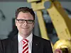 Abbildung Ralf Winkelmann von Fanuc, dunkler Anzug, gemusterte Krawatte, Brille. Steht vor Roboter