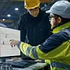 Zwei Mitarbeiter blicken auf Bildschirm mit Software (linker mit gelben Helm und rechter mit gelber Weste und weißem Helm)