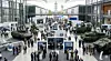 Übersichtsfoto aus der Vogelperspektive einer belebten Messehalle während der Verteidigungsmesse DSEI Germany in Hannover. Zu sehen sind diverse Stände mit Militärfahrzeugen, Radarsystemen, Drohnen und Bildschirmen. Fachpublikum in Anzügen und Militärunterform bewegt sich zwischen den Exponaten.