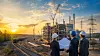 Ingenieure und Geschäftsleute prüfen Pläne vor einer Industriebaustelle mit Kränen, Bahngleisen und Energieinfrastruktur.