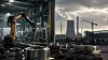 Symbolbild einer industriellen Fertigung mit Roboterarm, Metallbauteilen, Energieinfrastruktur und Baustelle im Hintergrund.