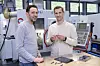 Zwei Männer stehen in einer Werkstatt neben einer CNC-Maschine und halten ein Bauteil in der Hand.