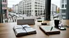 Holzschreibtisch in einem modernen Büro vor einem großen Panoramafenster mit Blick auf eine Stadtstraße; darauf liegen ein gebündelter Aktenstapel, ein Notizbuch mit Stift, eine Kaffeetasse und eine Messingwaage.